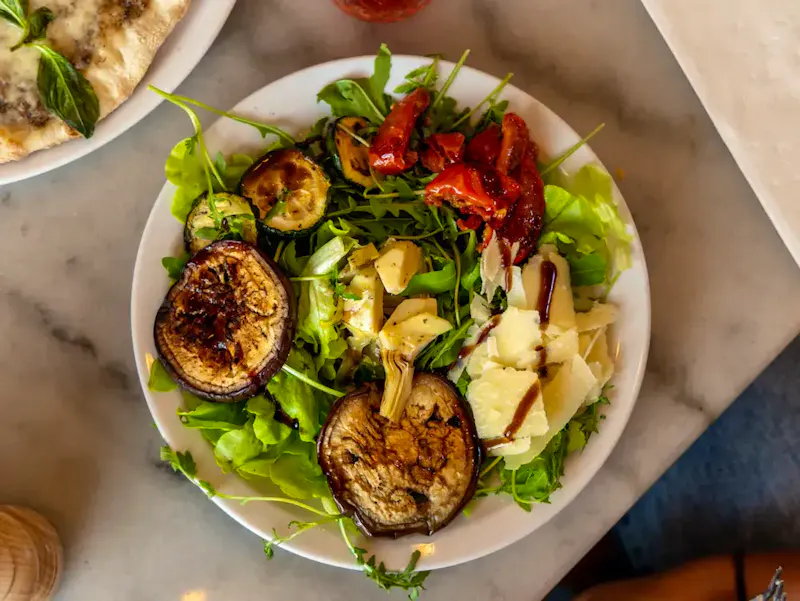 Assiette d'Antipasto di legumi du restaurant Ti Amo : un mélange de salade verte, cœurs d'artichauts, fines tranches de parmesan, aubergines et courgettes grillées, agrémenté de tomates confites savoureuses.