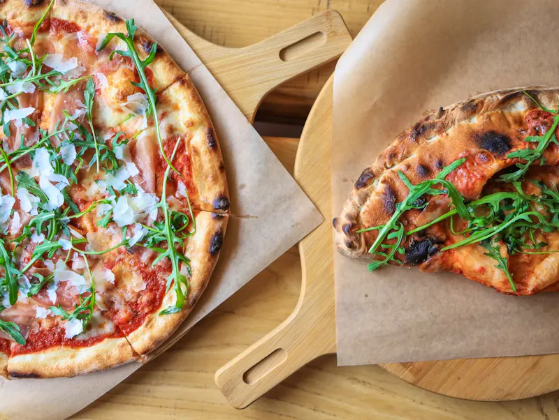 Image of La Vendetta Freshly baked pizzas topped with prosciutto, arugula, and cheese, served on wooden boards. | Photo by Valeria Boltneva, on https://www.pexels.com/@valeriya?utm_source=CentralApp&utm_medium=referral