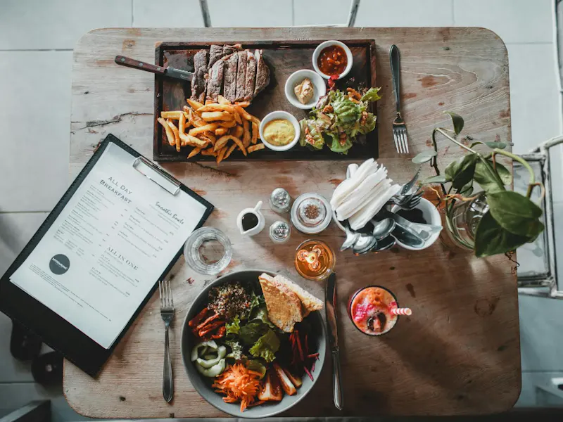 Top view of wooden table with salad bowl and fresh drink arranged with tray of appetizing steak and french fries near menu in cozy cafe | Photo by ROMAN ODINTSOV, on https://www.pexels.com/@roman-odintsov?utm_source=centralapp&utm_medium=referral
