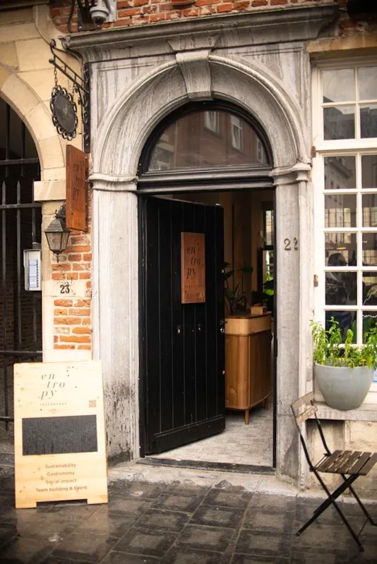 Vue de l'entrée du restaurant Entropy à Bruxelles. Une grande porte noire entrouverte est nichée sous une arche en pierre grise classique. À gauche, un panneau de bois incliné présente le logo "Entropy" et les piliers de l'établissement : Durabilité, Gastronomie, Impact social et Événements. La façade en briques rouges, une lanterne ancienne et une petite chaise pliante en bois sur le trottoir créent une atmosphère accueillante et authentique